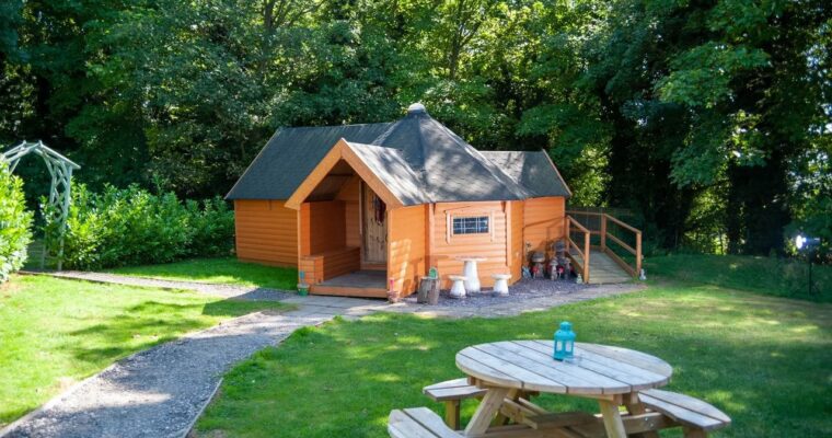Picnic bench and small wooden glamping hut, surrounded by trees at Kit's Coty