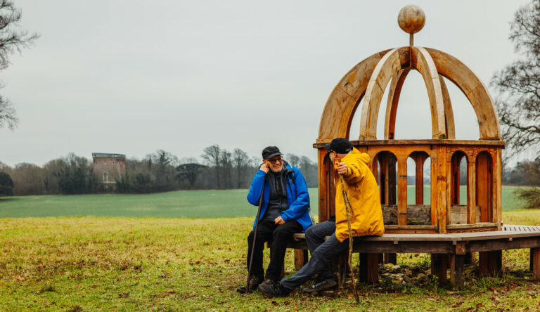 Two people with raincoats sitting on Monumenta Romana sculpture bench.