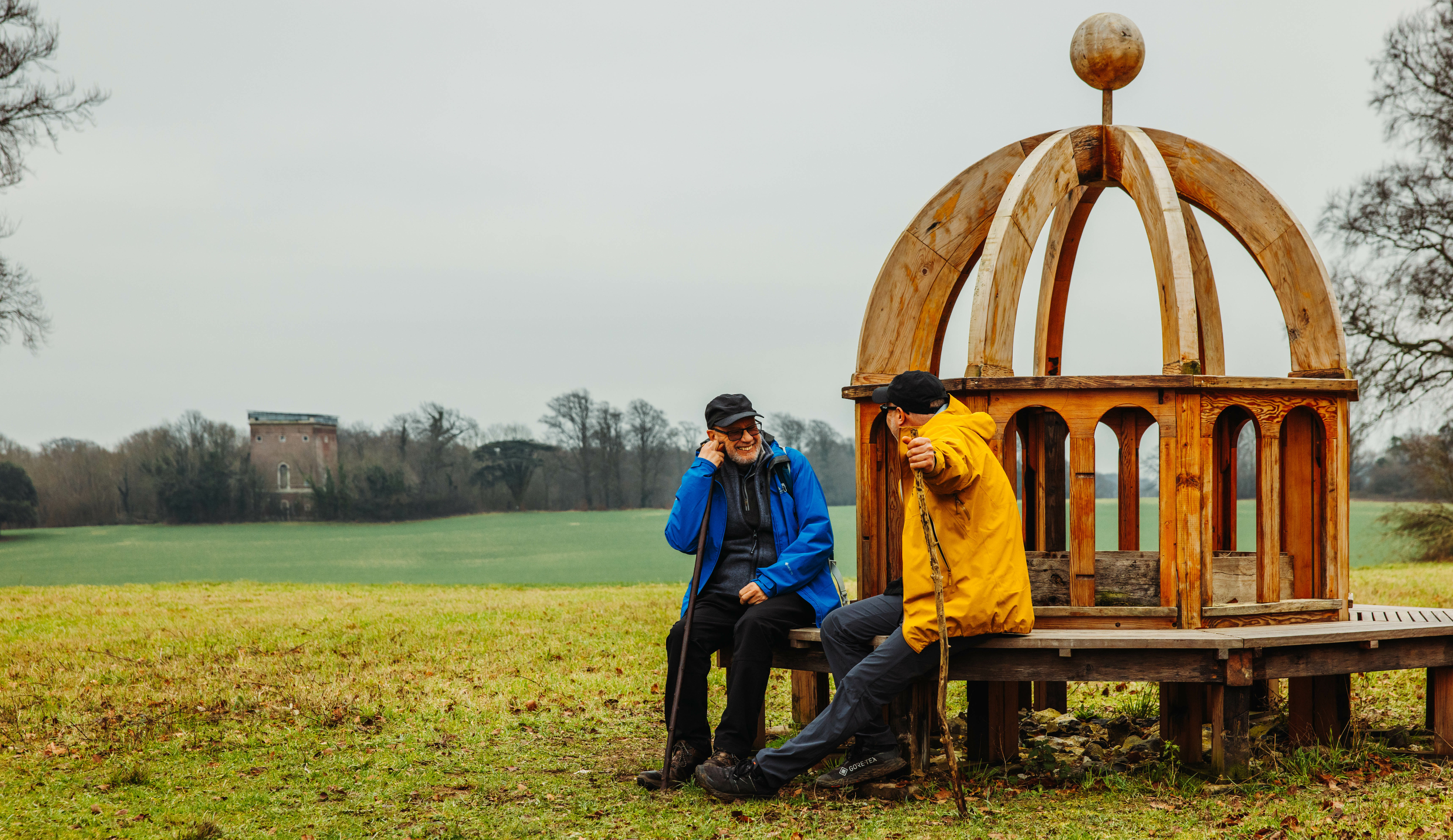Two people with raincoats sitting on Monumenta Romana sculpture bench.