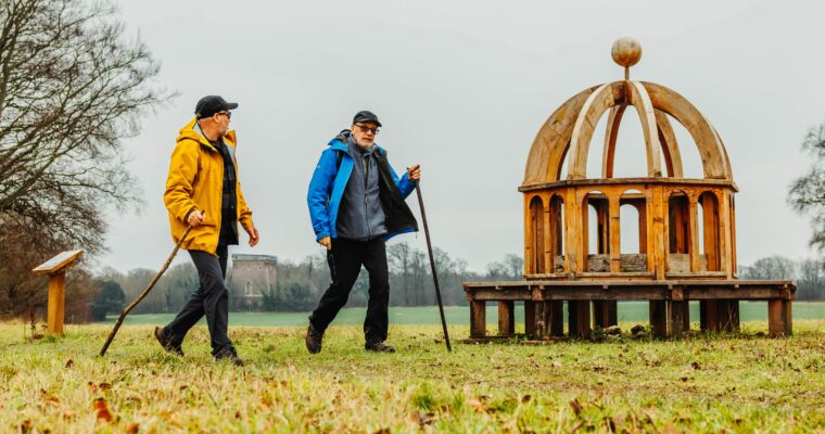 Two people walking on grassland near Monumenta Romana sculpture.