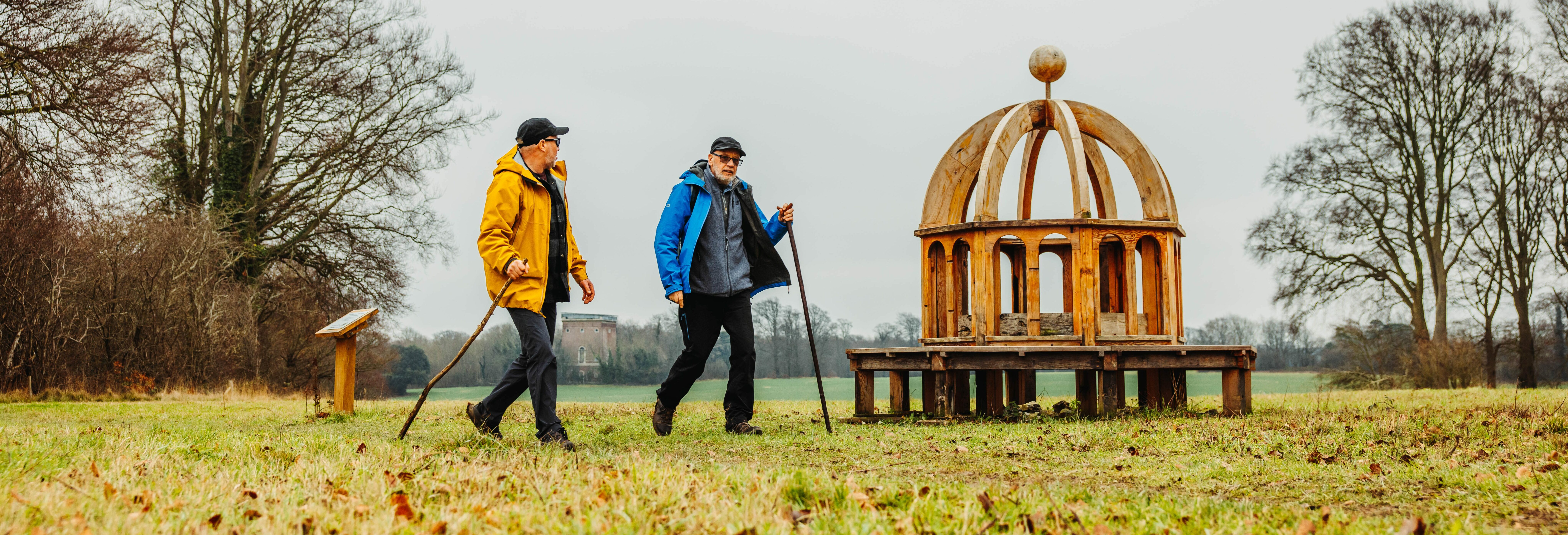 Two people walking on grassland near Monumenta Romana sculpture.