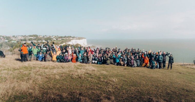 Big group of walkers, smiling and waving at camera for a group photo on grass with cliffs and sea in background.