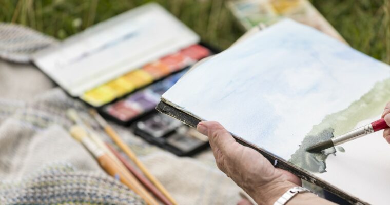 Close-up person holding a sketch pad, painting a picture in watercolours.