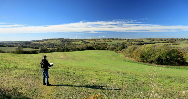 Green fields and trees in the distance, views across Stour Valley. Blue sky. Person looking at view with map in hand.