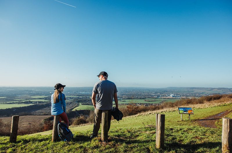 A man and woman perched on wooden posts, with a far reaching countryside view behind