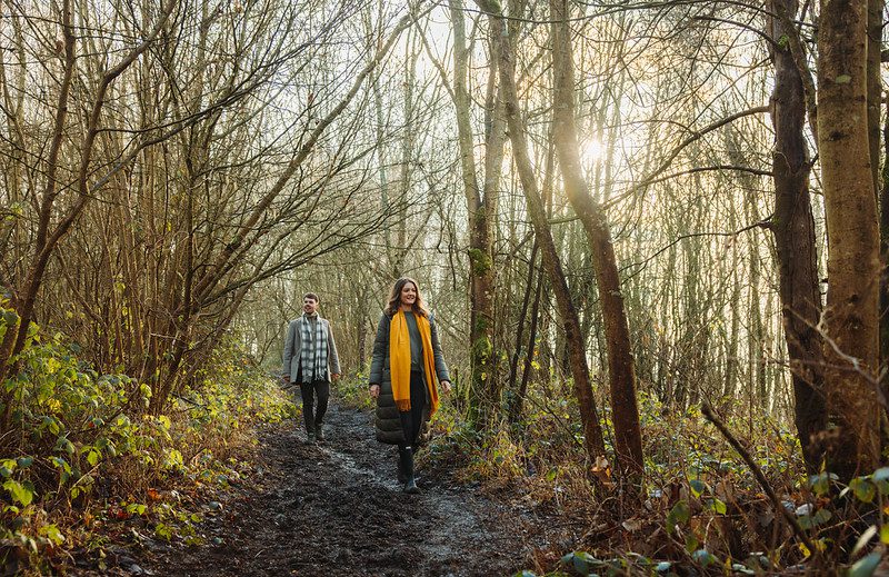 A man and woman wearing scarves walk along a muddy path through a wood