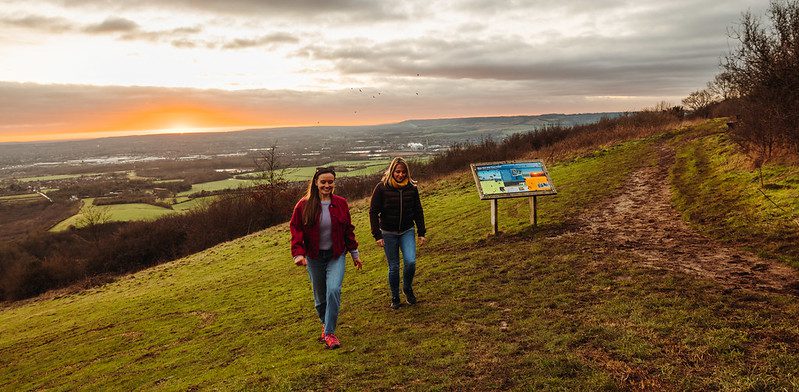 Two women walk away from a countryside sign with the sunsetting in the distance behind