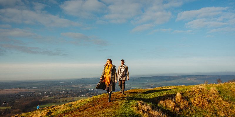 A man and woman walk atop a hill with far reaching panoramic views behind