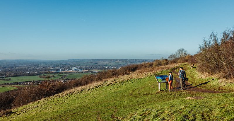 A man and woman stand atop a hill looking at a sign and the panoramic views