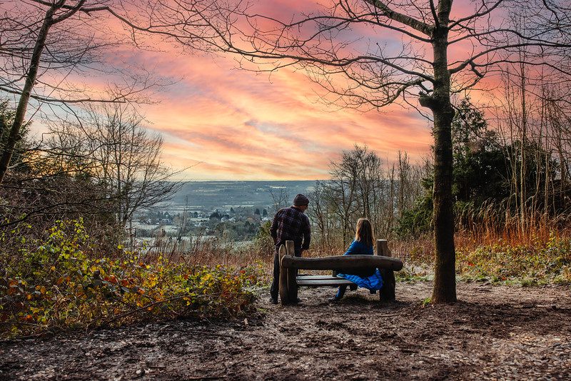 A woman sits on a bench next to a standing man, admiring view of pink sunset over far reaching countryside views