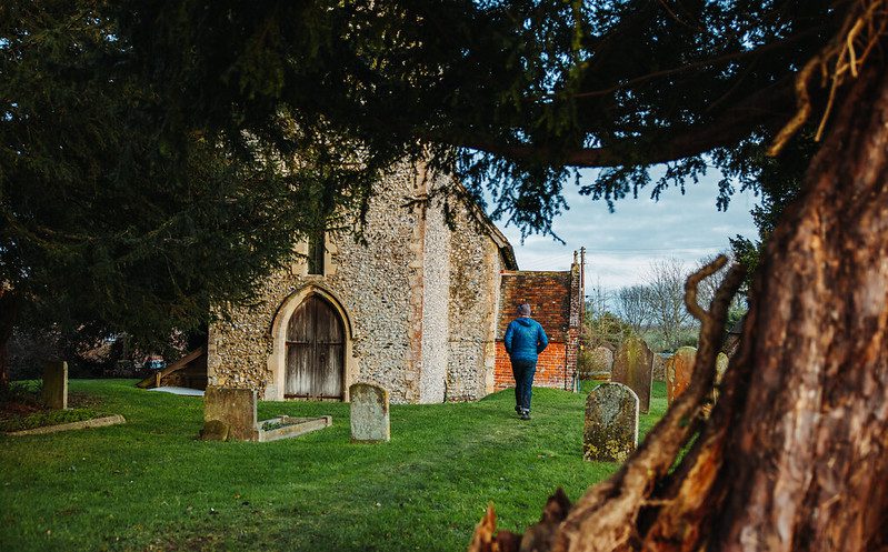 Man in blue coat walks towards a quaint church through the churchyard with gravestones