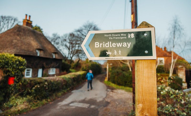 Close up of bridleway sign with thatched cottage and man walking down country lane in the background