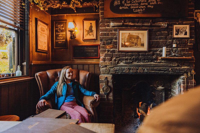 A smiling woman sits in a leather chair by a pub fire