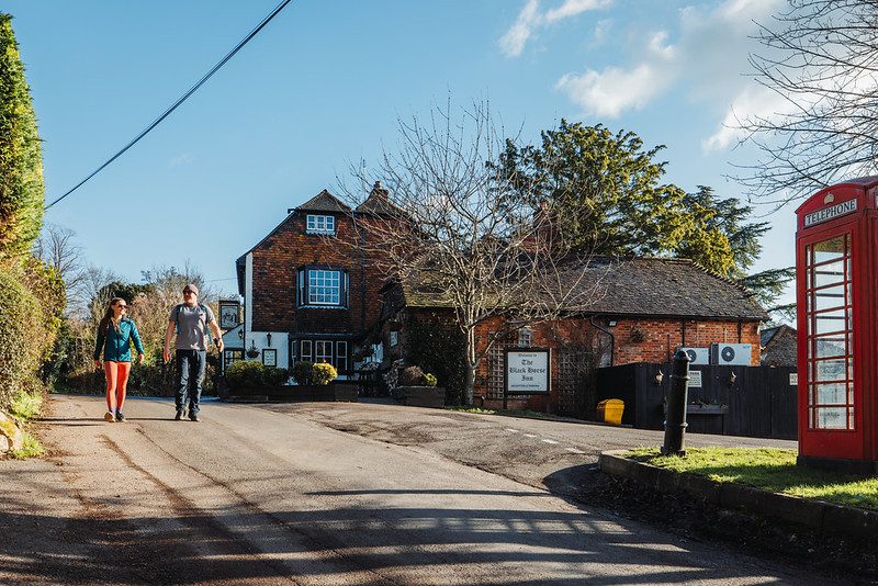 A man and a woman walking on a country lane with a pub and old English phonebox