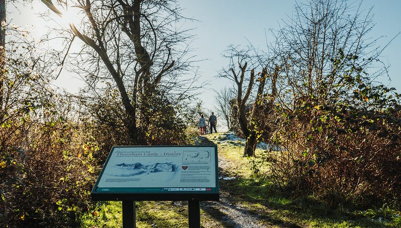 Sign for Thurnham Castle in the foreground with a couple looking at the remains behind