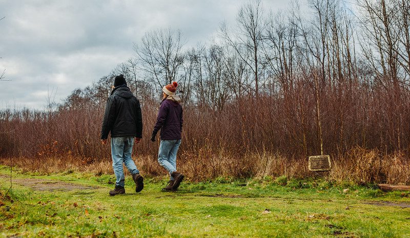 A couple in woolly hats and jeans walk through a field alongside a woodland in winter