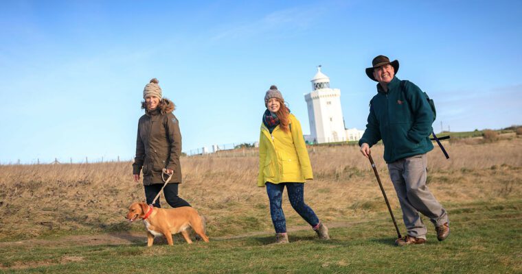 Three walkers with dog walking in front of white lighthouse