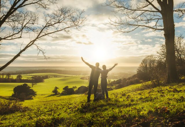 A couple with arms in the air looking at the sunset across a green landscape