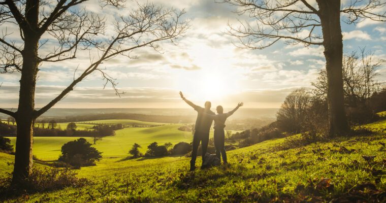 A couple with arms in the air looking at the sunset across a green landscape