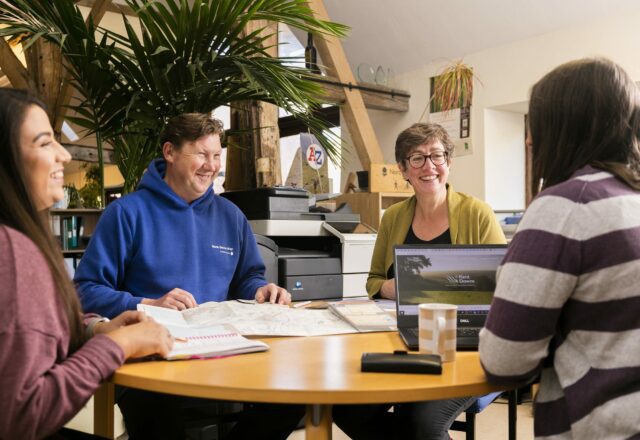 Four smiling people sitting around a table in an office with laptops and notepads