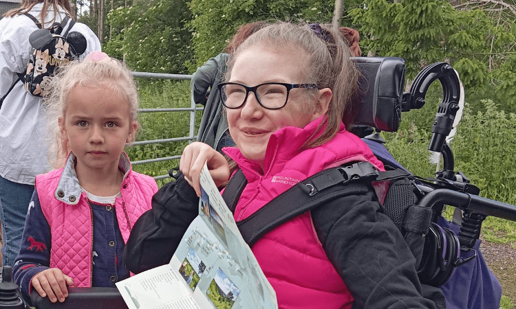 A young girl in a wheelchair holds up a North Downs Way National Trail map. A younger girl stands next to her. 