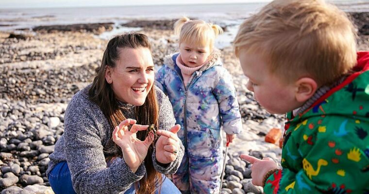 Adult and two small children, looking as crab on seashore. Pebble beach and sea in background.