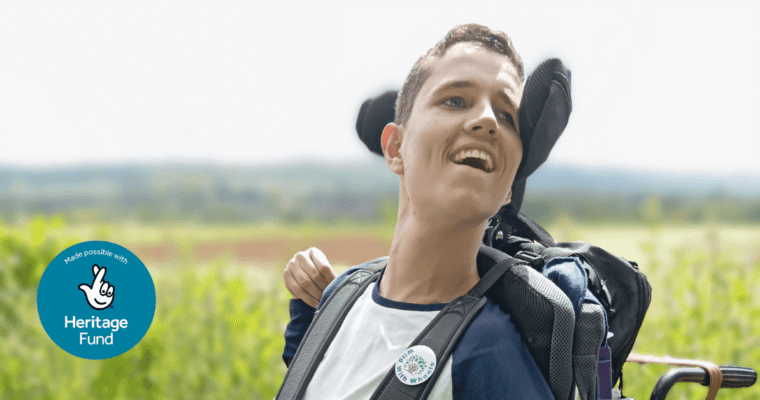 A young man in a wheelchair is in the foreground of the image. The background is trees, fields and foliage. This is part of the North Downs Way National Trail.
