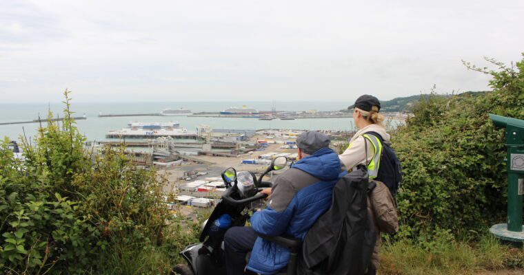 A man in a mobility scooter overlooks the port of Dover. A woman stands next to him. There are boats and vegetation in the landscape.