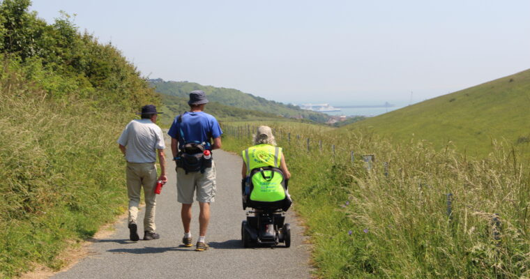 Three individuals with their back to the camera follow a flat path through grassland. One person is using an electric mobility aid. In the distance is white cliffs and the sea.