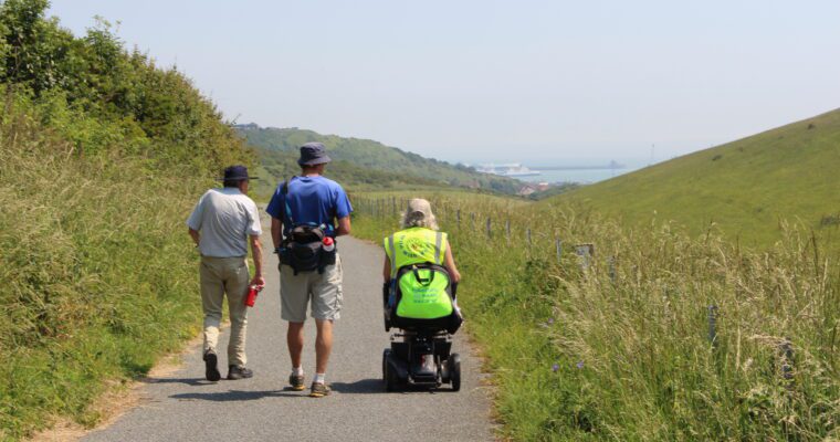 Three individuals with their back to the camera follow a flat path through grassland. One person is using an electric mobility aid. In the distance is white cliffs and the sea.