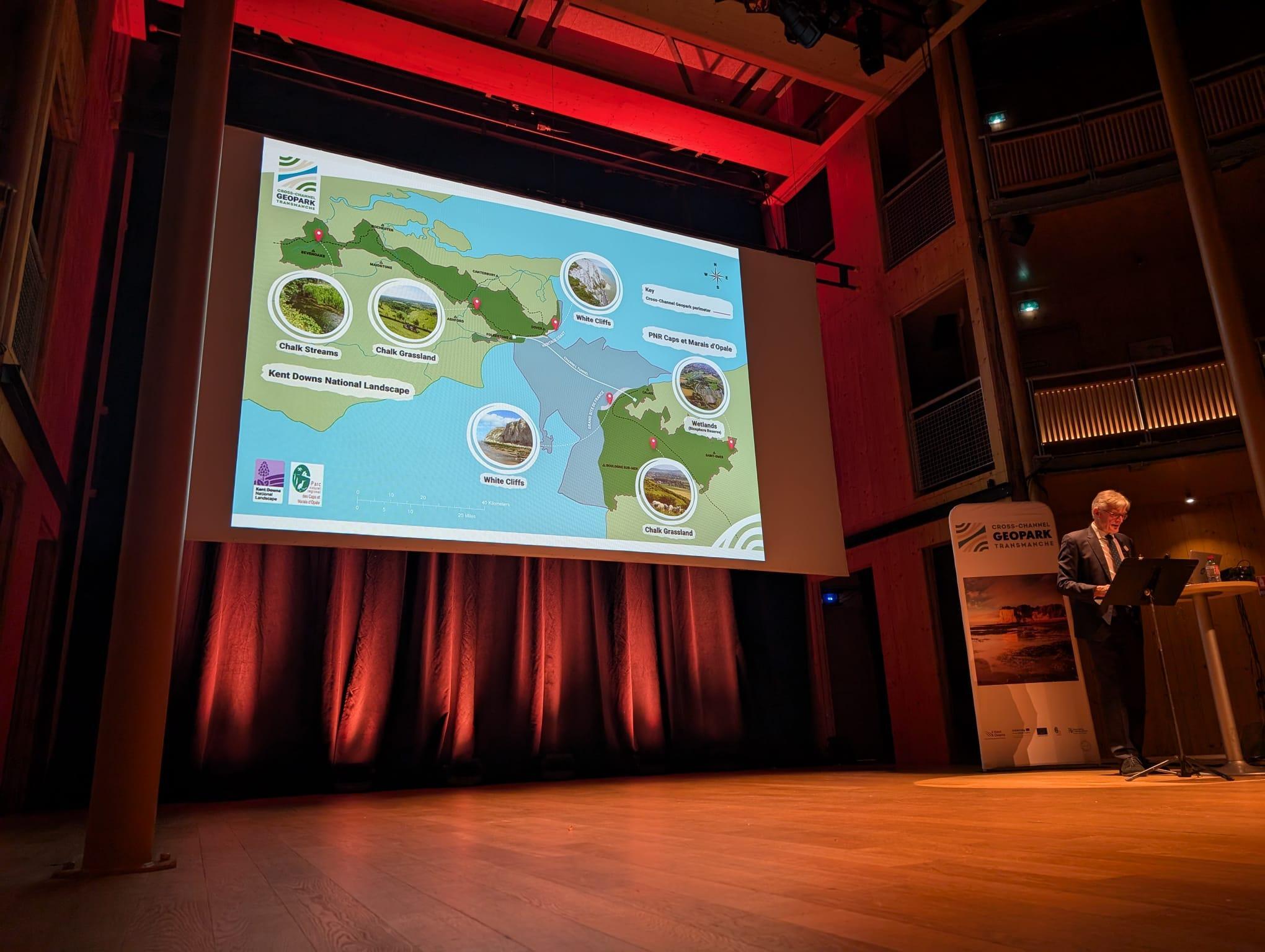 The image shows Nick Crane presenting the geopark boundary, with a large map displayed in the background.