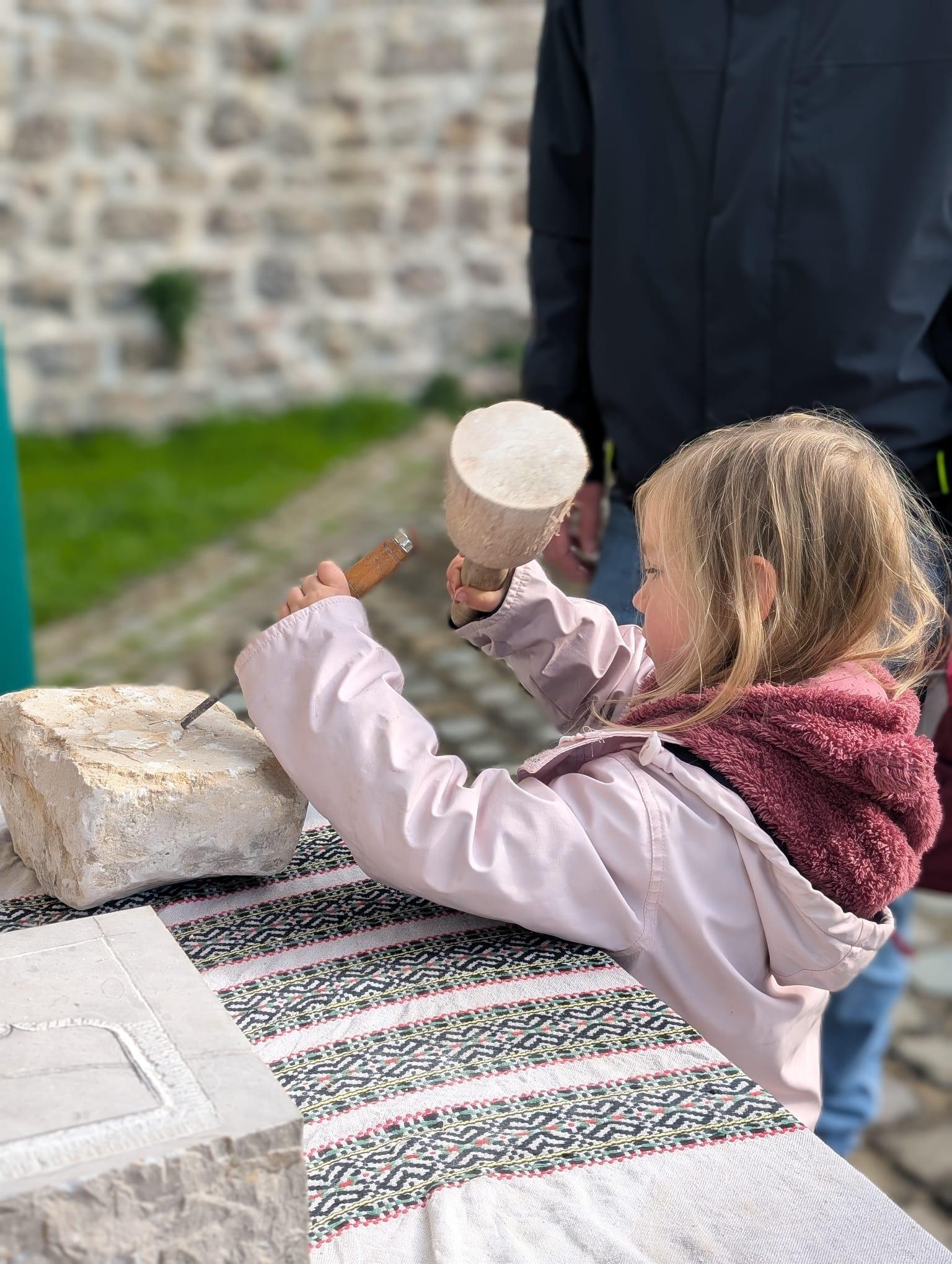 A young girl tries her hand at being a stonemason, carefully chiselling a piece of stone.