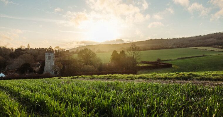 The sun begins to set behind the historic St Peter & St Paul Church in Trottiscliffe, casting a warm glow over the surrounding fields. The church stands against a soft, colorful sky with shades of orange and pink. The gently rolling fields in the foreground are bathed in the golden light of the evening, creating a peaceful, rural scene.