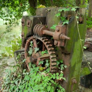 Ruins of Dartford Mill featuring old, rusted machinery among the stone walls and overgrown plants by the riverside.