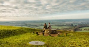 Two people sit on a circular stone structure atop a grassy hill, gazing over a vast landscape of fields and distant towns beneath a cloudy sky.