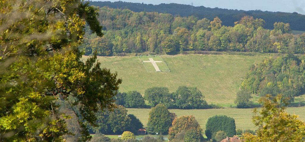 A large white cross stands prominently on a hillside, contrasting with the surrounding green grass. In the foreground, trees with green and yellow leaves frame the scene, suggesting an autumn setting. Below the hill, fields and a small cluster of buildings form a village or town. The background features a dense forest with a mix of green and autumnal hues. The cross, a striking focal point, may hold cultural, historical, or religious significance.