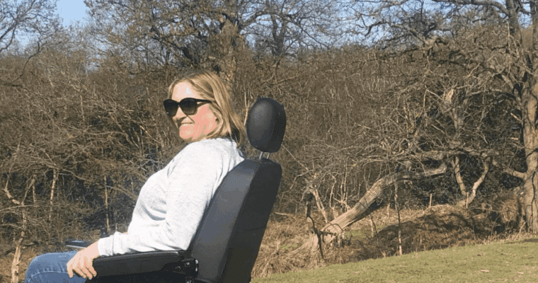 Woman with sunglasses sitting in a wheelchair on a grassy area with trees in the background.
