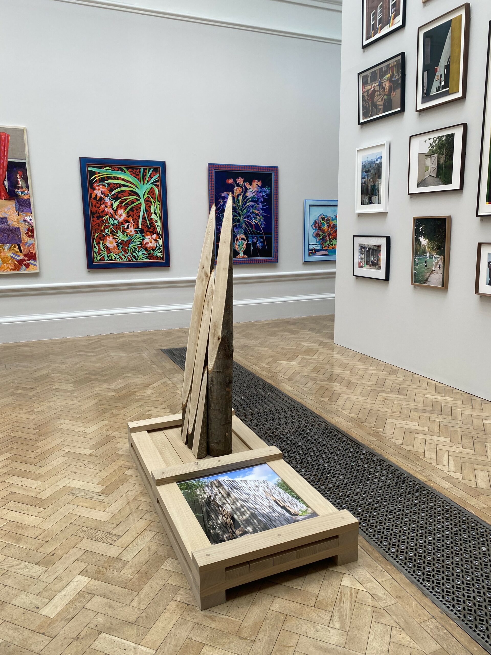 A well-lit art gallery with parquet flooring features colorful framed paintings and photographs on the walls; a central wooden rack holds large, unmounted artworks beside a floor-level metal grate, suggesting an organized space for exhibition setup or storage.