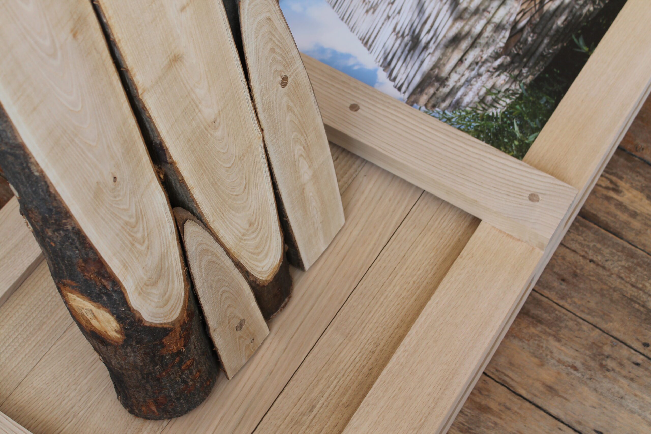 Close-up of “Coppice Oratory” highlighting its raw textures: upright logs with bark intact contrast against a smooth, framed wooden panel, while a backdrop photograph of a tree canopy and blue sky deepens the connection to woodland origins.