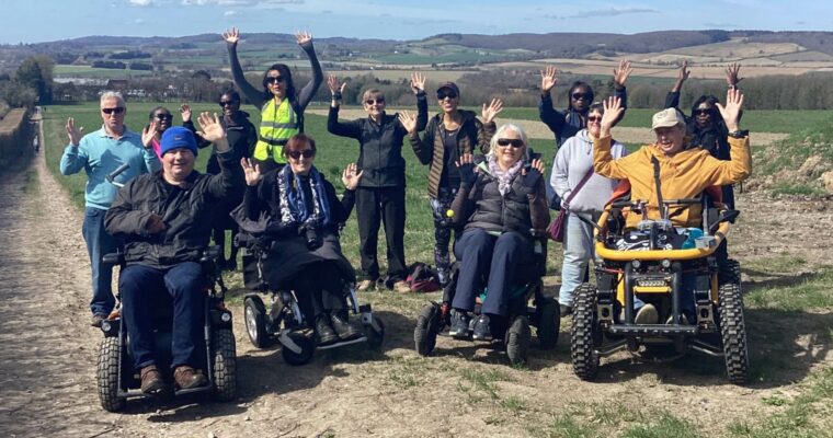 A group of people in wheelchairs raising their hands while on a trail outdoors.