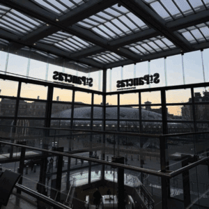 A view from inside St Pancras International station, showing its glass ceiling and reflections of the station name across tall windows and steel arches.