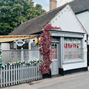 A charming white tea house named The Honey Pot, with heart-themed windows, a climbing vine and a vintage-style sign, set behind a picket fence and closed for the day.