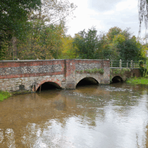 A red-and-stone arched bridge stretches across a quiet river, framed by autumn trees and soft reflections in the water.