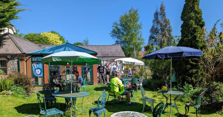 A sunny garden scene with green metal tables and chairs on a grassy lawn.