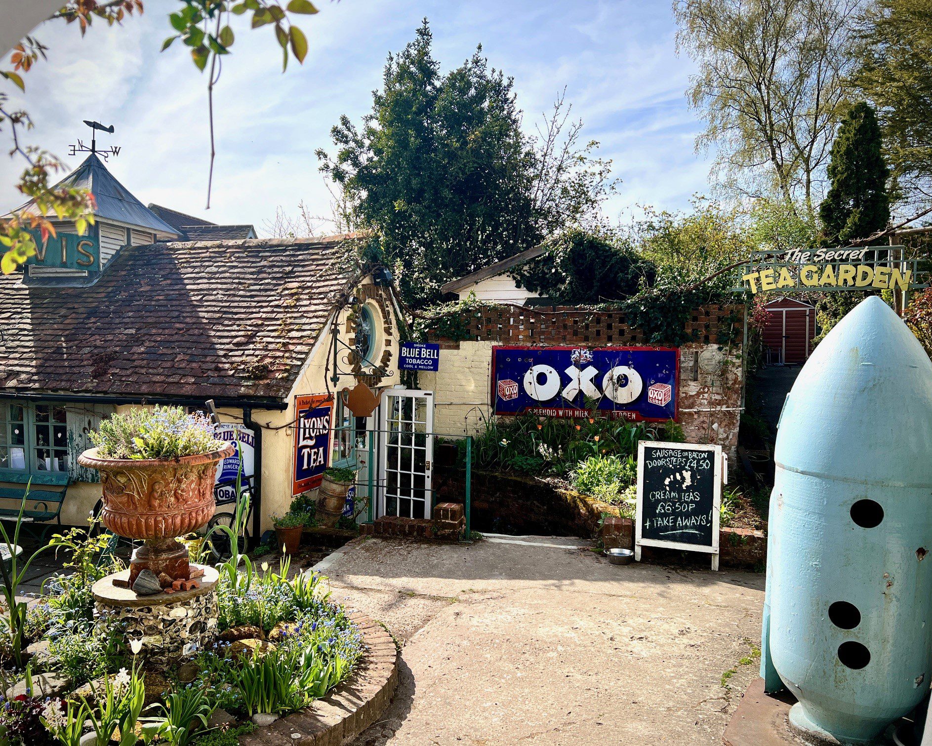 A quaint tea garden with a rustic building, vintage signs, and a large flower-filled planter