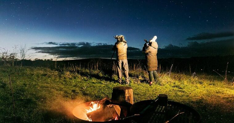 Two people standing looking at the night sky, through a telescope, in a field with camp fire behind them.
