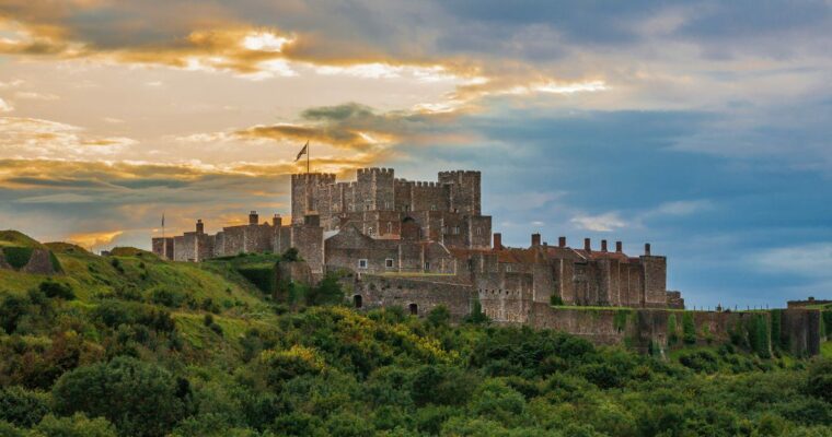 Dover Castle, a large medieval fortress with stone walls and towers, sits atop a green hill surrounded by dense vegetation under a dramatic sky with clouds and patches of golden sunlight.