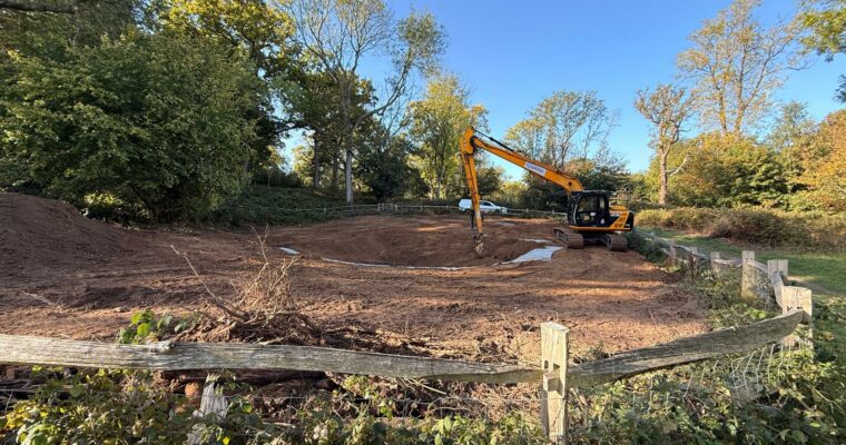 Adding the Bentonite pond liner. Excavator working on a construction site in a woodland area, shaping earth for a pond or landscape project. The scene includes a fenced-off area with soil mounds, trees with autumn foliage, and a clear blue sky.