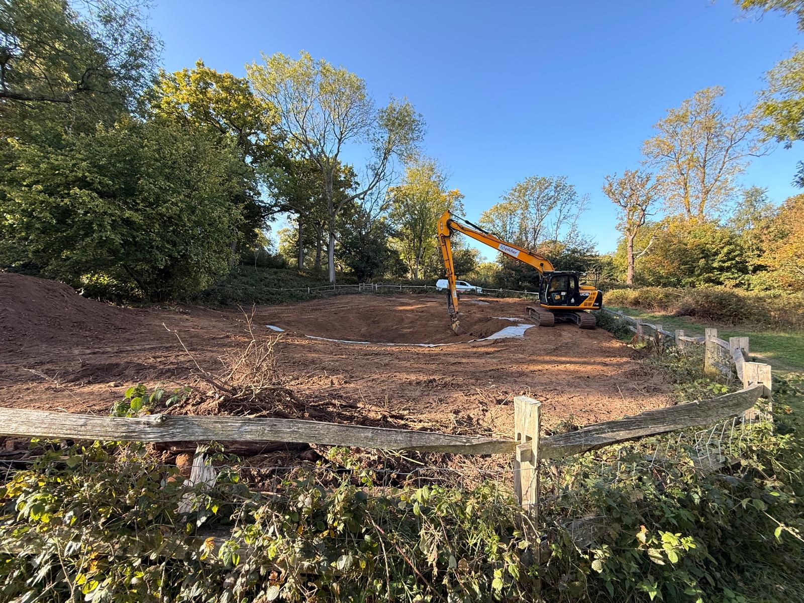 Adding the Bentonite pond liner. Excavator working on a construction site in a woodland area, shaping earth for a pond or landscape project. The scene includes a fenced-off area with soil mounds, trees with autumn foliage, and a clear blue sky.