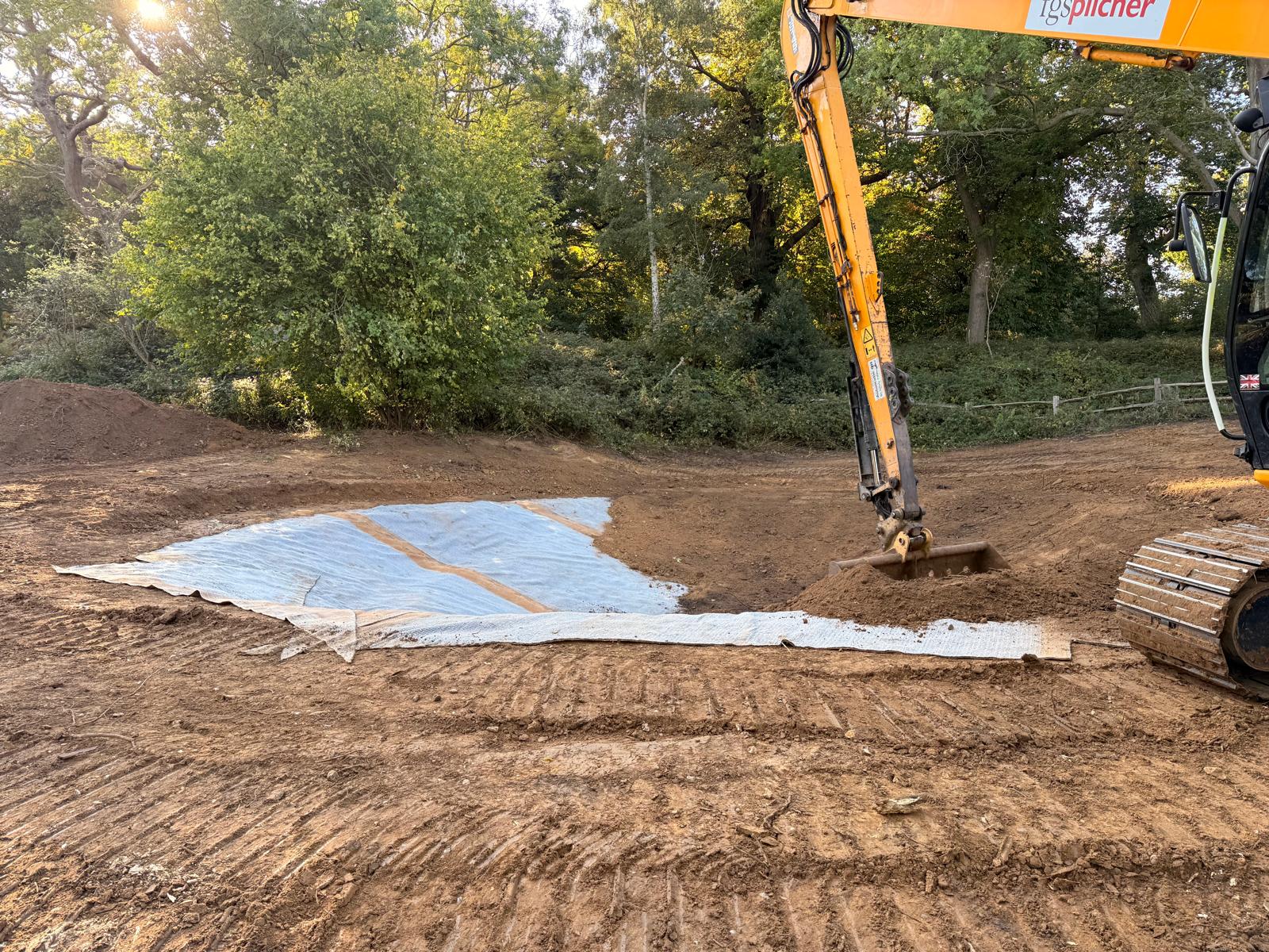 Adding the Bentonite pond liner. Excavator placing bentonite liner on a prepared soil surface during pond construction in a woodland clearing, with trees and vegetation in the background under natural daylight.